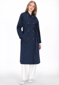 Marineblauwe trenchcoat met dubbele rij knopen, ceintuur en grote zijzakken. Glad, waterbestendig materiaal, knielengte ontwerp.