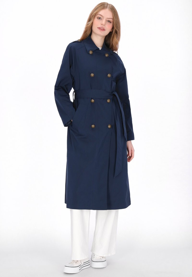 Marineblauwe trenchcoat met dubbele rij knopen, ceintuur en grote zijzakken. Glad, waterbestendig materiaal, knielengte ontwerp.
