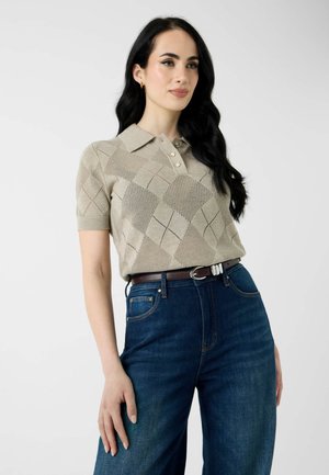 Femme aux longs cheveux noirs portant un pull beige à manches courtes avec col et un jean bleu taille haute avec une ceinture marron.
