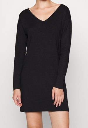 Kobieta w czarnej, dzianinowej sukience z długim rękawem i dekoltem w serek, stojąca na tle jasnej, jednolitej ściany, z rękami swobodnie opuszczonymi wzdłuż ciała.