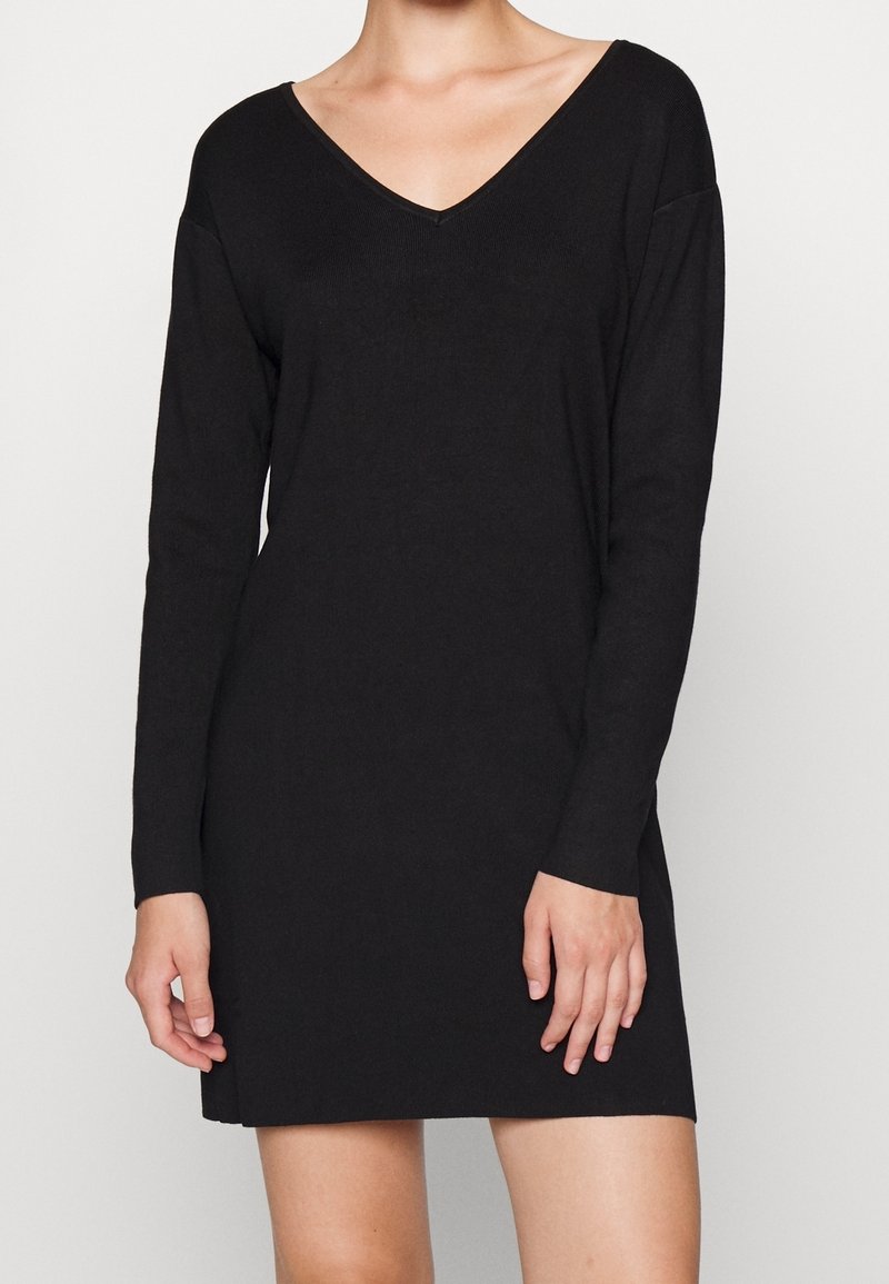 Femme portant une robe en maille noire à manches longues et col en V, debout devant un fond clair uni, les mains détendues le long du corps.