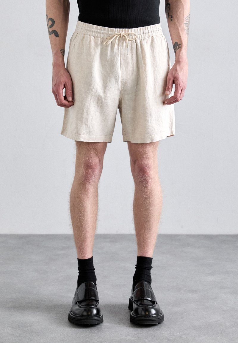 Shorts en lin beige avec une coupe décontractée, une taille élastique et un cordon de serrage. Présentent des fentes sur les côtés et une texture légère et respirante.