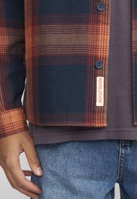 Camisa a cuadros en navy oscuro, naranja y beige; cuenta con botones redondos y una etiqueta que dice "REVOLUCIÓN." Llevada sobre una camiseta gris oscura y jeans.
