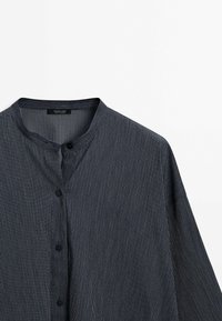 Chemise gris foncé à boutons avec fines rayures verticales et col rond à bande sur fond blanc.