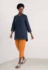 Marine linnen tuniek met een kraaghalslijn, driekwartmouwen en zijsplitsingen, gecombineerd met oranje losse broeken en grijze sandalen.