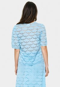 Top in pizzo di colore azzurro chiaro con un motivo scallopato, piccole maniche a sbuffo e una vestibilità comoda. Il materiale traslucido mette in evidenza il design a strati.
