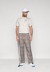 Vyriškas golfo aprangos derinys, kuriame yra šviesiai smėlio spalvos smulkiai raštuotas polo marškinėlis su žaliu logotipu ir rudos spalvos languoti kelnės, papildytos baltais bateliais ir balta kepure.