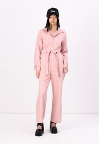 Macacão rosa com fecho na frente, gola com volantes, bolsos laterais e cintura amarrada. Combinado com sapatos plataforma pretos e um boné preto.