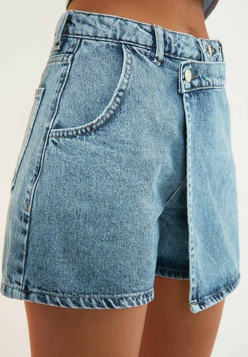 Short en jean bleu clair avec un rabat frontal superposé, passants de ceinture, fermeture par bouton argenté et poches latérales à la taille et sur le haut des cuisses d’une personne.