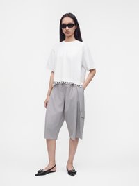Γυναίκα που φοράει μαύρα γυαλιά ηλίου, λευκό κοντομάνικο τοπ με τελείωμα πομ πομ, φαρδιά γκρι σορτς μέχρι το γόνατο και μαύρα μυτερά μπαλαρίνια με αγκράφες.