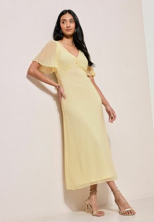 Frau in einem blassgelben Midikleid mit plissierten Flügelärmeln, Knöpfen am Oberteil und goldenen, schnürbaren High Heels, die vor einer hellen Wand posiert.