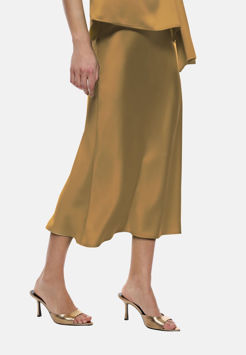Gonna midi in satin color senape, caratterizzata da un design fluido. Abbinata a sandali con tacco aperto dorati con tacco strutturato.