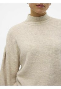 Gros plan d'une personne portant un pull à col roulé côtelé beige, montrant le haut du torse et le cou.