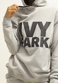 Szara bluza z kapturem z napisem "IVY PARK" w kolorze czarnym, wyposażona w sznurki przy kapturem i ściągacze przy rękawach. Miękka tekstura materiału; luźny krój.