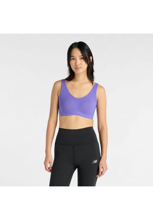 Vrouw die een paarse sportbeha en zwarte hoge taille legging draagt, staand tegen een effen witte achtergrond.