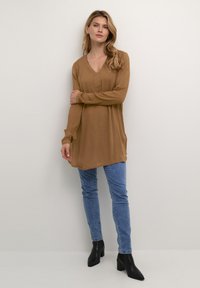 Tunica a maniche lunghe e scollo a V in una calda tonalità marrone, realizzata in tessuto morbido. Abbinata a jeans blu aderenti e stivaletti neri con punta.