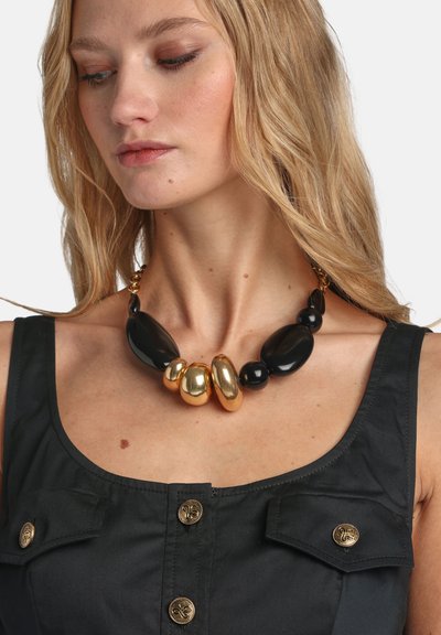 Collier audacieux composé de perles ovales lisses noires et brillantes dorées, de tailles variées. Haut en tissu texturé avec des boutons décoratifs dorés visibles.