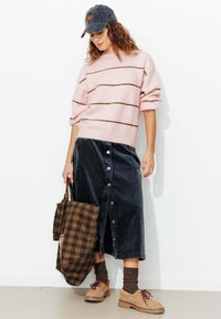 Femme portant un pull à rayures roses, une jupe foncée boutonnée, des chaussettes marron, des chaussures beiges, une casquette en jean, tenant un grand sac à carreaux marron.