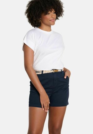 Kobieta o kręconych włosach ubrana w białą koszulkę, granatowe szorty i pleciony pasek, stojąca z jedną ręką w kieszeni.