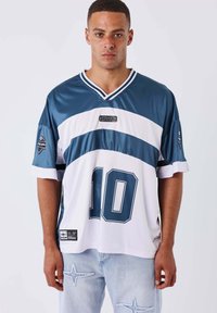 Giovane uomo che indossa una maglietta sportiva blu e bianca con il numero 10, abbinata a jeans azzurri chiari con toppe a forma di stella.