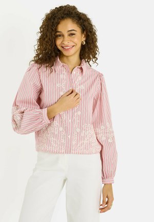 Mujer sonriente con cabello rizado, vistiendo una blusa de rayas rosas y blancas con bordado y pantalones blancos, posando frente a un fondo liso.