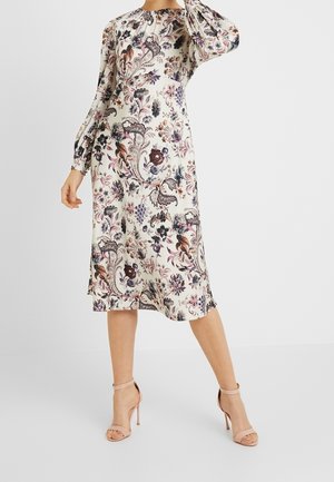 Sukienka w kwiaty w kształcie litery A z długimi rękawami, w kremowej bazie z fioletowymi i czerwonymi wzorami oraz okrągłym dekoltem. Do tego neutralne szpilki.