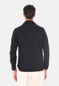 Camicia nera con bottoni, dalla texture morbida, colletto a bavero e spacchi sulle maniche; indossata sopra pantaloni chiari.