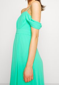 Mintunvihreä, laineileva mekko, jossa on spagettiolkaimet ja olkapäät paljastavat hihoja. Ominaisuuksiin kuuluu muotoonommeltu yläosa ja laskostettu hame rentoa siluettia varten.