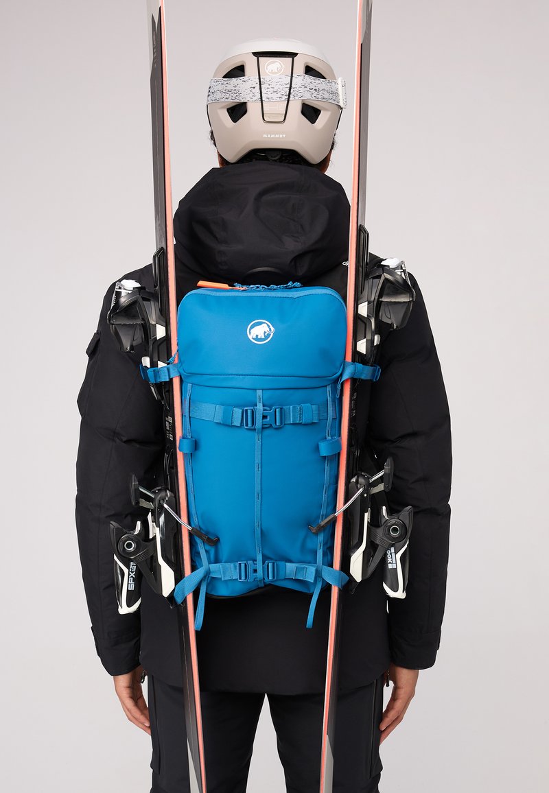 Blauwe rugzak met verstelbare banden, slank ontwerp, voorvak en meerdere bevestigingspunten. Op beide zijden gemonteerde ski's zichtbaar.