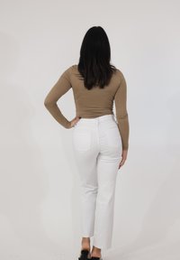 Kobieta o długich ciemnych włosach, ubrana w beżową bluzkę z długim rękawem, białe spodnie i czarne szpilki z odkrytymi piętami, stojąca z jedną ręką na biodrze.