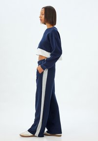 Marineblauem, tailliertem Sweatshirt mit grauem Saum, kombiniert mit marineblauen, weit geschnittenen Hosen mit grauen Seitenstreifen und Taschen. Das Material wirkt weich und strukturiert.