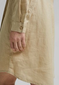 Gros plan d'une main reposant sur le côté portant une robe en lin beige à manches longues avec détail de bouton de manchette.