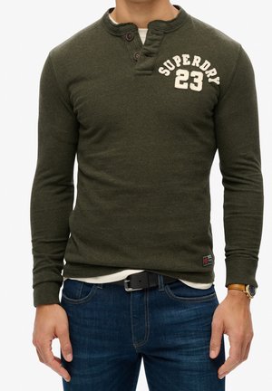 Olivgrünes Langarmshirt mit Henley-Ausschnitt, ausgestattet mit drei Knöpfen und dem Schriftzug "SUPERDRY 23" in Creme, kombiniert mit dunkelblauen Jeans.