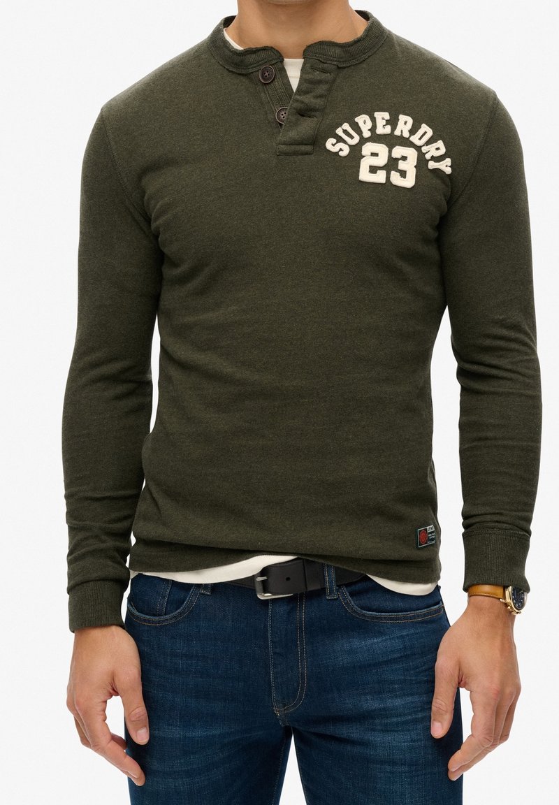 Olivengrøn langærmet skjorte med henley-udskæring, der har tre knapper og "SUPERDRY 23" i creme, kombineret med mørkeblå jeans.