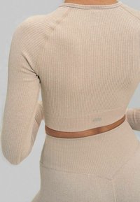 Top beige de manga larga con textura acanalada, diseño corto y silueta ajustada; cuenta con costuras reforzadas y una pequeña etiqueta con el logo.