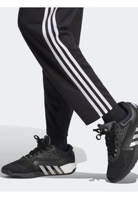 Schwarze Sporthosen mit drei weißen Streifen an den Seiten. Schwarze Turnschuhe mit silbernen Akzenten, Netzpaneelen und Schnürsenkeln.