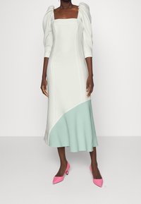 Robe blanche à manches ballon, décolleté carré et ourlet asymétrique vert menthe ; associée à des escarpins roses à bouts pointus.