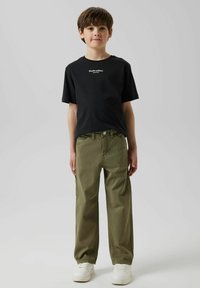 Zwarte katoenen T-shirt met de tekst "Studio editie"; olijfgroene wijdvallende broek; witte sneakers; effen achtergrond.