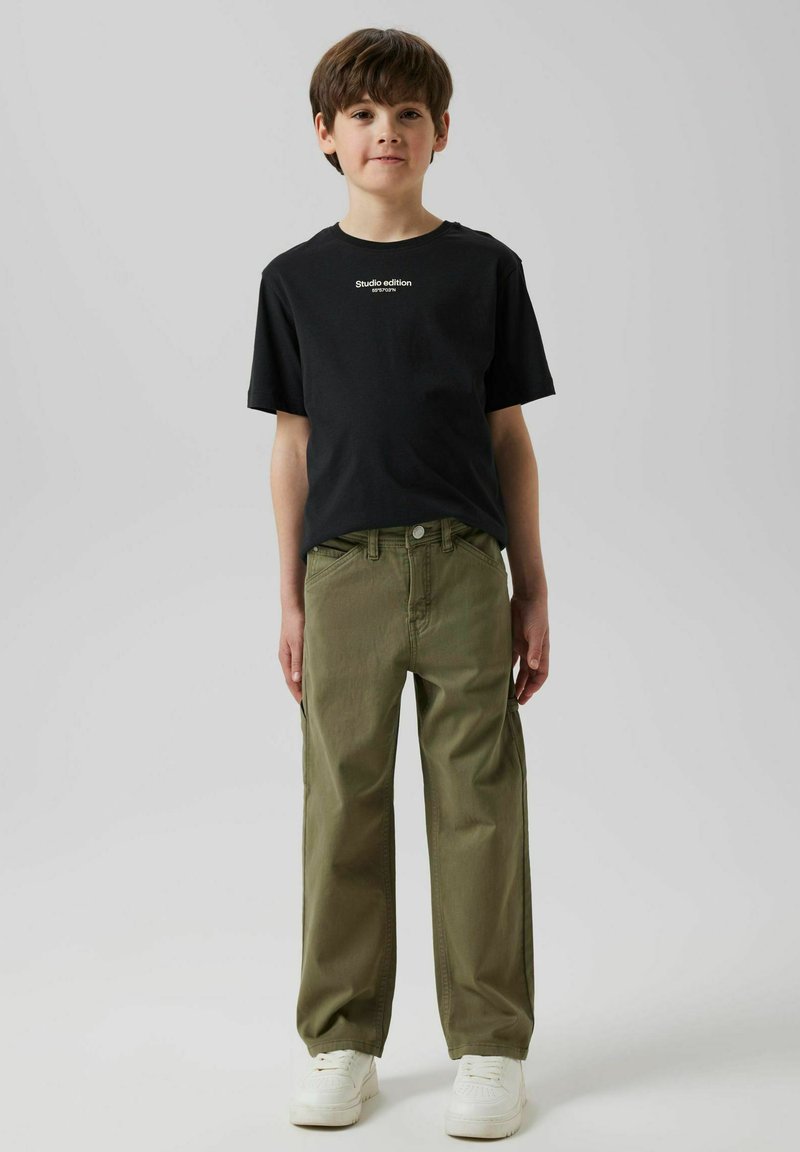 Zwarte katoenen T-shirt met de tekst "Studio editie"; olijfgroene wijdvallende broek; witte sneakers; effen achtergrond.