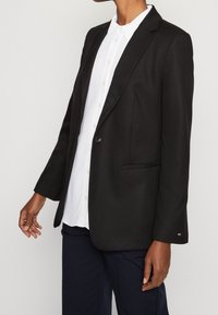 Svart kavaj i slätt tyg med en knapp, slagkrage och sidoficka. Bärs över en vit skjorta med krage.