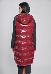 Czerwona błyszcząca kamizelka puchowa z kapturem, z pikowanym wzorem i poziomymi paskami. Widok z tyłu ukazuje błyszczącą fakturę i smukłą sylwetkę.