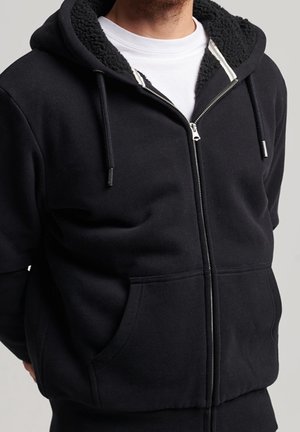 Mann trägt einen schwarzen Kapuzenpullover mit Reißverschluss, fleecegefütterter Kapuze und Kordeln über einem weißen T-Shirt mit Rundhalsausschnitt, steht vor einem einfarbigen Hintergrund.