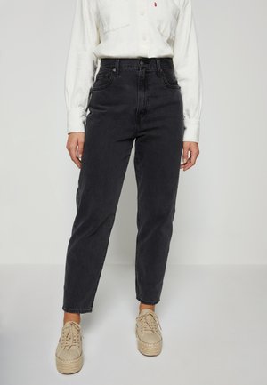 Person trägt schwarze hoch taillierte Jeans, weißes langärmliges Hemd eingesteckt und beige Schnür-Plateauschuhe und steht vor einem einfarbigen Hintergrund.