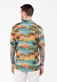 Homme avec des bras tatoués portant une chemise hawaïenne colorée ornée de palmiers, de vues sur l'océan et de fleurs oranges, associée à un pantalon blanc.