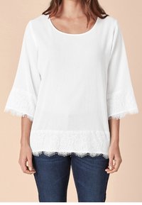 Chemisier blanc avec un col rond et des manches cloche en dentelle trois quarts. La bordure en dentelle en bas ajoute de la texture et des détails. Porté avec un jean foncé.