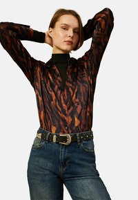 Koszula w brązowy i czarny wzór zwierzęcy z zapięciem na guziki, zestawiona z czarnym paskiem z srebrnymi akcentami i niebieskimi dżinsami.