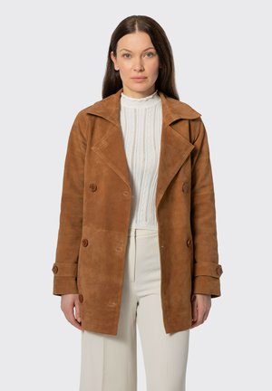 Vrouw draagt een bruine suède double-breasted jas over een witte, gestructureerde top met hoge hals en crèmekleurige broek, staand tegen een effen achtergrond.