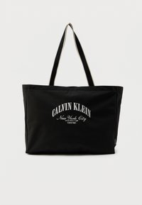 Μαύρη τσάντα tote από ανθεκτικό υλικό με την επιγραφή «CALVIN KLEIN New York City» σε λευκή γραμματοσειρά· διπλές λαβές και ορθογώνιο σχήμα.