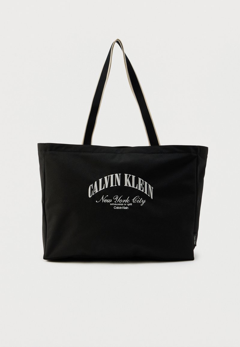 Μαύρη τσάντα tote από ανθεκτικό υλικό με την επιγραφή «CALVIN KLEIN New York City» σε λευκή γραμματοσειρά· διπλές λαβές και ορθογώνιο σχήμα.