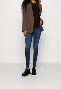 Hnědý blazer, černý top s krajkovým lemem, modré skinny džíny a černé nazouvací boty. Blazer má dvě kapsy a volný střih.